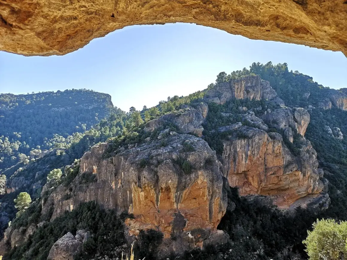 Paisaje rocoso y boscoso visto a través de un arco natural. La ruta del agua de Chelva se revela en la distancia.
