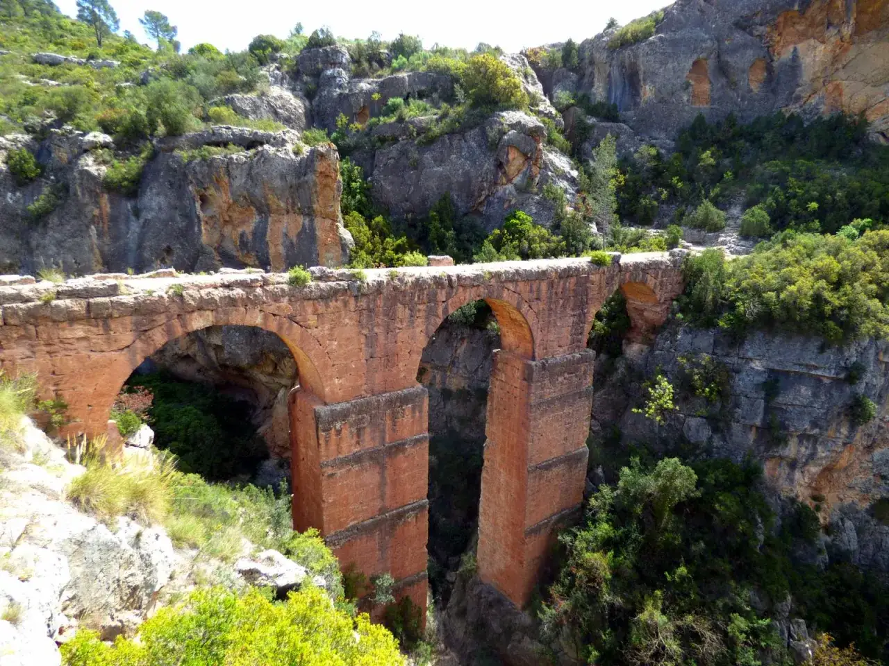Antiguo acueducto de piedra, parte de la ruta del agua de Chelva, cruza un barranco rodeado de vegetación y rocas.