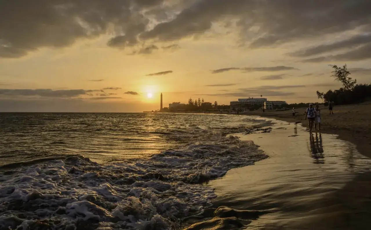 Atardecer en las dunas de Maspalomas, con el mar acariciando la arena y siluetas de personas paseando.